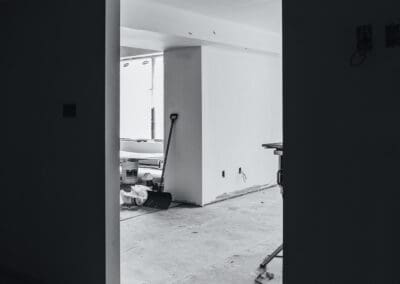 Black and white image of an unfinished room interior seen through a doorway, showing a shovel, paint buckets, and construction debris on a concrete floor.