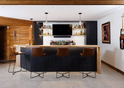 Modern home bar featuring a light wood countertop, black cabinetry, multiple liquor shelves, a wall-mounted TV, and four brown leather bar stools. The space includes wood paneling, a concrete floor, and a wall-mounted electric guitar with artwork.