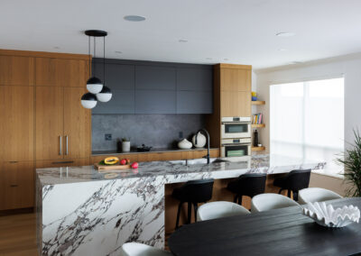 Modern kitchen with large white and grey marble waterfall island, light wood cabinetry, dark grey upper cabinets, and black pendant lights. A dining table with chairs is visible in the foreground.