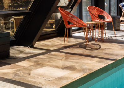 Modern indoor-outdoor pool area with two orange woven lounge chairs, a small matching table, natural stone flooring, and a large sloped glass roof reflecting light.