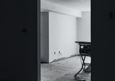 Black and white image of an unfinished room viewed through a doorway, showing white walls, exposed wiring, a concrete floor, and a power tool on a stand.