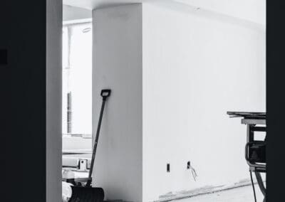Black and white photo of an unfinished room interior during renovation, showing white walls, exposed wiring, and a shovel leaning against a wall.
