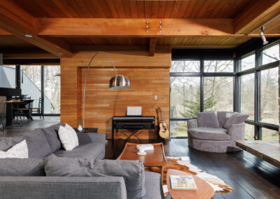 A modern rustic living room featuring a gray sectional sofa, dark wood floors, a natural wood plank wall, and an exposed wood beam ceiling. Large windows offer views of a winter forest, while a digital piano, acoustic guitar, cowhide rug, and arc floor lamp complete the cozy, artistic space.