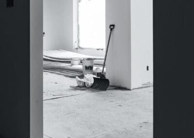 Black and white image of an interior room under renovation, showing a shovel, construction materials, and covered floor.