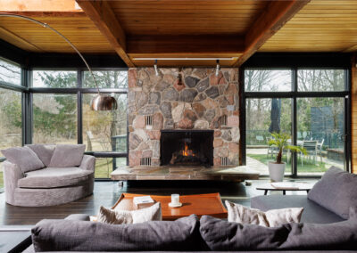 Spacious modern living room with a grand natural stone fireplace, exposed wooden ceiling beams, dark hardwood floors, and panoramic full-height windows overlooking a lush outdoor landscape and patio. Contemporary grey sofas and an architectural curved floor lamp furnish the sophisticated space.