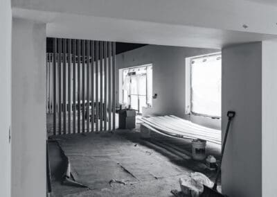 Black and white photo of an unfinished commercial interior space during renovation, featuring exposed drywall, windows covered in plastic, a slatted partition wall, and construction materials like stacked trim boards and tools on a protective floor covering.