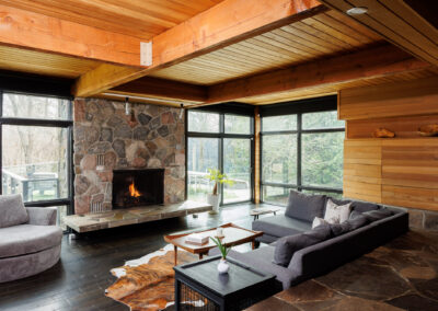 A modern rustic living room featuring a large stone fireplace, wood beam ceiling, expansive windows with nature views, a gray sectional sofa, and a cowhide rug on dark wood and flagstone flooring.