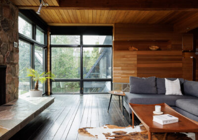 Modern living room with large windows, stone fireplace, wood paneling, grey sectional sofa, and dark hardwood floors.