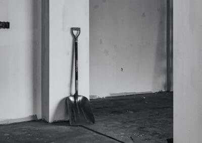 A Garant D-handle shovel leans against a white column in a grimy, unfinished room, rendered in grayscale.