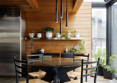 Modern dining area with natural wood paneling, a glass-top table, black chairs with straw seats, and large windows with greenery.