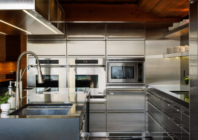 A modern kitchen featuring integrated stainless steel appliances, custom metal cabinetry, a dark stone island with a sink, a warm wooden plank ceiling with beams, and a dark flagstone floor.