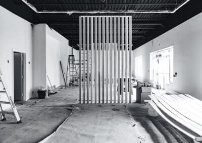 Black and white image of an office interior under construction, featuring a modern vertical slat wood partition, exposed industrial ceiling, unfinished white walls, ladders, and building materials.