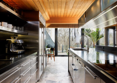 Modern kitchen with stainless steel appliances, black countertops, wooden ceiling, and large windows overlooking a natural forest. Features an espresso machine, integrated sink, and flagstone floor.