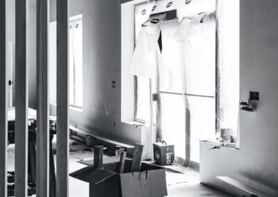 Black and white photo of an interior construction site with unfinished walls, large windows draped in protective plastic, raw wooden architectural slats, and various building materials on the floor, including a cardboard box with wood scraps and stacked primed boards.