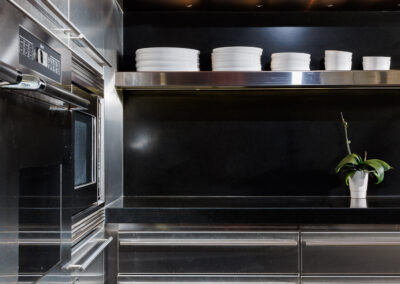 Modern kitchen with stainless steel cabinets, built-in oven, and open shelves displaying white dinnerware, complemented by a black countertop and a wooden ceiling.