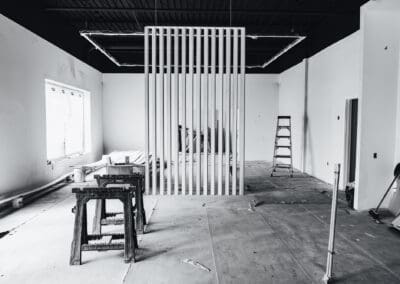 Black and white photo of an empty commercial space under construction with a modern slat partition, saw horses, and a ladder.