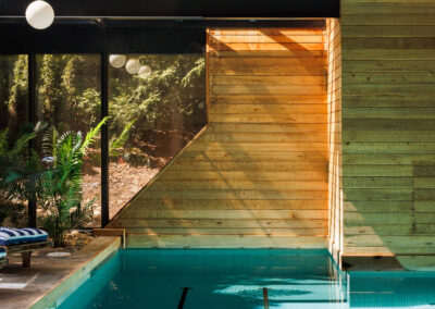 Modern indoor swimming pool with wood paneling, large windows showcasing forest views, and dappled sunlight creating shadows on the turquoise tiled water.