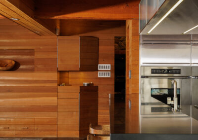Modern kitchen interior featuring rich wooden paneling, exposed ceiling beams, flagstone flooring, stainless steel appliances, and a dark kitchen island with contemporary wood bar stools.