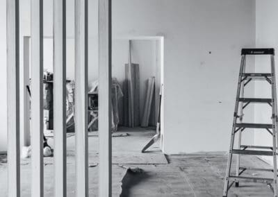 Black and white image of an interior space under construction, featuring a vertical wooden slatted partition in the foreground, an aluminum step ladder, and an unfinished floor and walls.