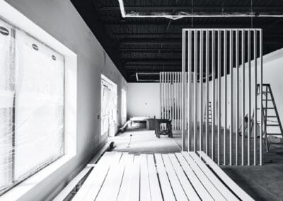 Black and white image of a modern interior construction site with white vertical slat walls, an exposed black ceiling, and protected windows, showing renovation in progress.