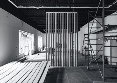 Black and white interior construction scene with a modern slat wall divider, scaffolding, and white trim boards on a concrete floor.