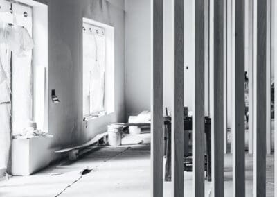 Black and white photo of an interior construction site with vertical wooden slats, unfinished concrete floor, and windows covered in protective plastic.