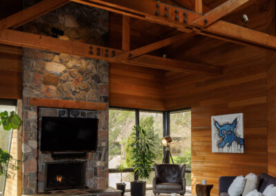 Rustic modern living room with a large stone fireplace, mounted TV, dark sectional sofa, wood-beamed vaulted ceiling, flagstone floor, and expansive windows.