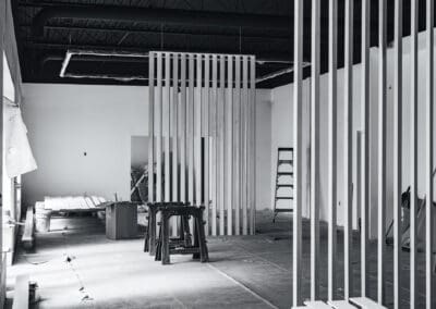 Black and white photo of a commercial interior under renovation, featuring newly installed vertical wooden slat room dividers, an exposed industrial black ceiling with HVAC ducts, and construction tools like saw horses and ladders on an unfinished floor.