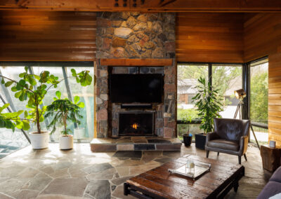 Modern rustic living room with large stone fireplace, TV, wood ceiling, flagstone floor, and indoor plants