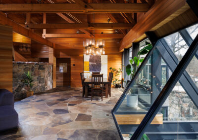 Rustic modern interior design with exposed wood beams, flagstone flooring, a wooden dining table, a multi-tier chandelier, and large angular glass windows