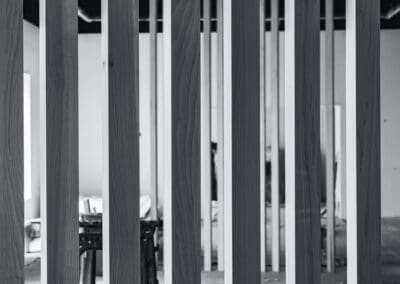 Black and white image of vertical wooden slats forming a modern room divider or architectural screen, with a blurred interior space visible behind.