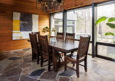 Modern rustic dining room with a dark wood table, six chairs, warm wood-paneled walls, an abstract painting, and large windows revealing a natural outdoor view.