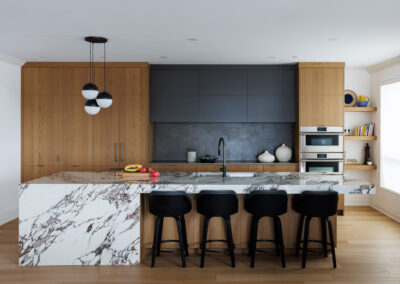 Modern kitchen with white marble waterfall island, light wood cabinetry, and dark gray upper cabinets. Features include black bar stools, stainless steel double oven, and spherical pendant lights.