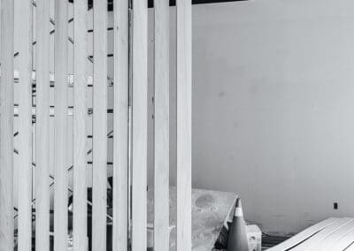 Black and white image of a modern wooden slat partition in an unfinished interior space, featuring a concrete floor, a white wall, and construction materials.