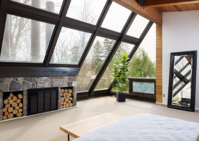 Modern bedroom with large sloped windows, a stone fireplace with firewood storage, and a fiddle leaf fig plant.