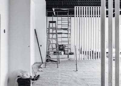 Black and white image of an unfinished commercial interior under construction, showing scaffolding, a ladder, exposed industrial ceiling, and modern vertical wooden room dividers.
