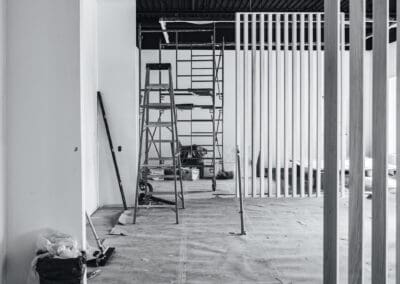 Black and white image of an interior construction site featuring scaffolding, a stepladder, and modern vertical slatted room dividers, indicating ongoing renovation.