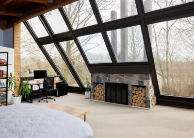 Modern bedroom with home office, large angled windows, stone fireplace, and a natural wooded view.