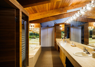 Luxurious bathroom with warm wood paneling, exposed ceiling beams, a long marble double vanity with sinks, large mirrors, and a built-in tub area.