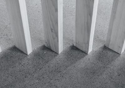 Vertical light wooden planks standing on a textured gray concrete floor, casting shadows in a minimalist grayscale composition.