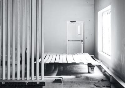 Black and white photo of an interior construction site with a wooden slat partition, white planks laid on a concrete floor, an emergency exit door, and a window.