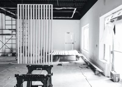 Black and white image of an interior commercial space under renovation, featuring scaffolding, a modern wooden slat wall partition, exposed ceiling, and windows protected with plastic sheeting.