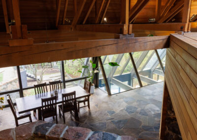 High-angle view of a modern home interior featuring exposed wooden beams, a wooden dining table, and large geometric windows with a stone floor.