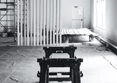 Black and white image of an unfinished interior space with sawhorses, a slat room divider, and construction elements.