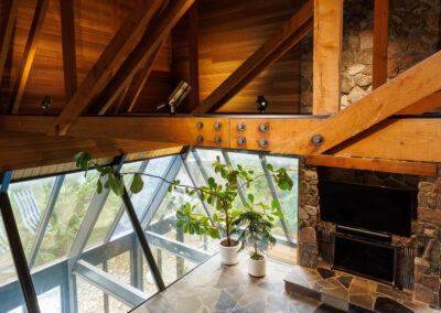 Modern rustic living room featuring exposed wooden beams, a stone fireplace, large angled glass windows, and flagstone flooring.