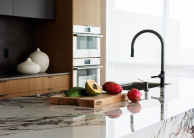 Modern kitchen island with white and reddish-brown veined marble countertop, featuring a wooden cutting board with papaya and dragon fruit, a matte black faucet, and contemporary wood and dark gray cabinets in the background.