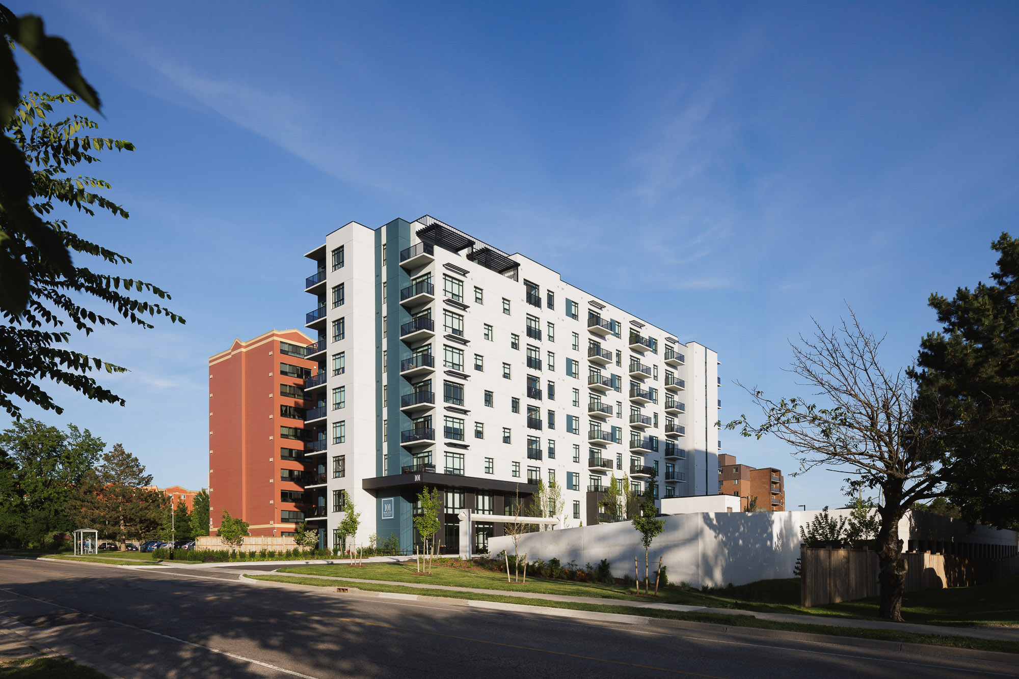 Modern white apartment building with balconies and teal accents under a clear blue sky, viewed from a street.