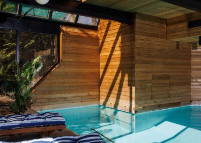 Luxury indoor swimming pool with wooden paneling, large glass windows, two wooden lounge chairs with blue and white striped cushions, and spherical pendant lights.
