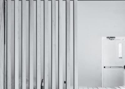 Black and white image of a modern wooden slat room divider in an industrial interior space under construction, with an emergency exit door in the background.
