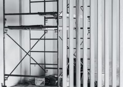 Black and white image of an indoor construction site with a metal scaffolding unit and a partially installed vertical wooden slat wall or room divider, showcasing ongoing interior renovation.